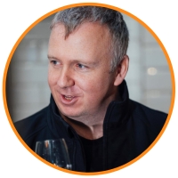 Grey haired man wearing a dark coloured and collared top talking and looking to the side. Joe McGirr CEO & Founder of Boatyard Distillery