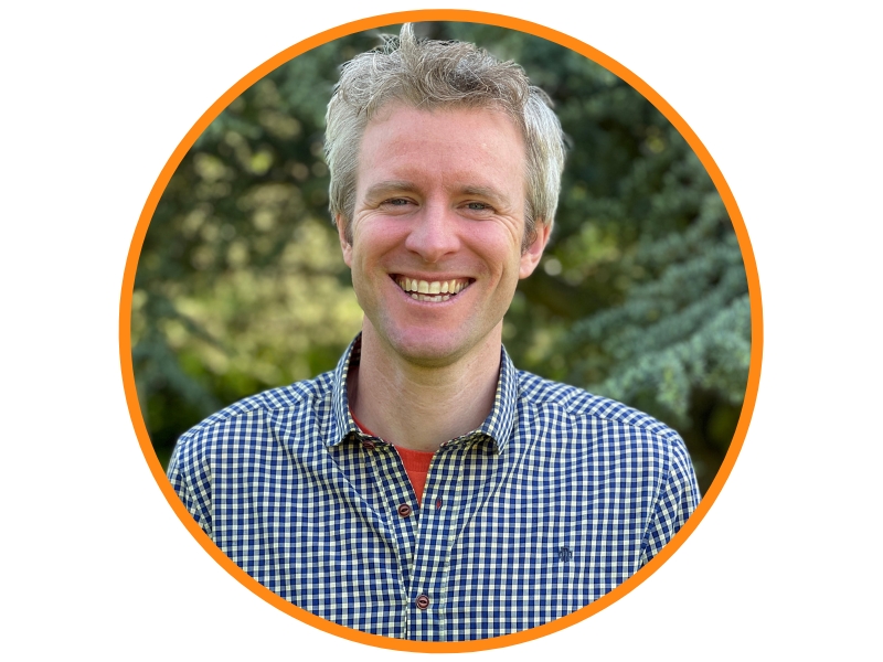 Grey haired man smiling in front of trees and wearing a checked navy and white shirt and red vest