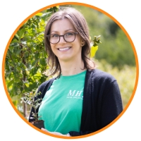 Brown haired woman standing outside in front of a green natural area, smiling while holding a small tree, wearing glasses and a green t-shirt