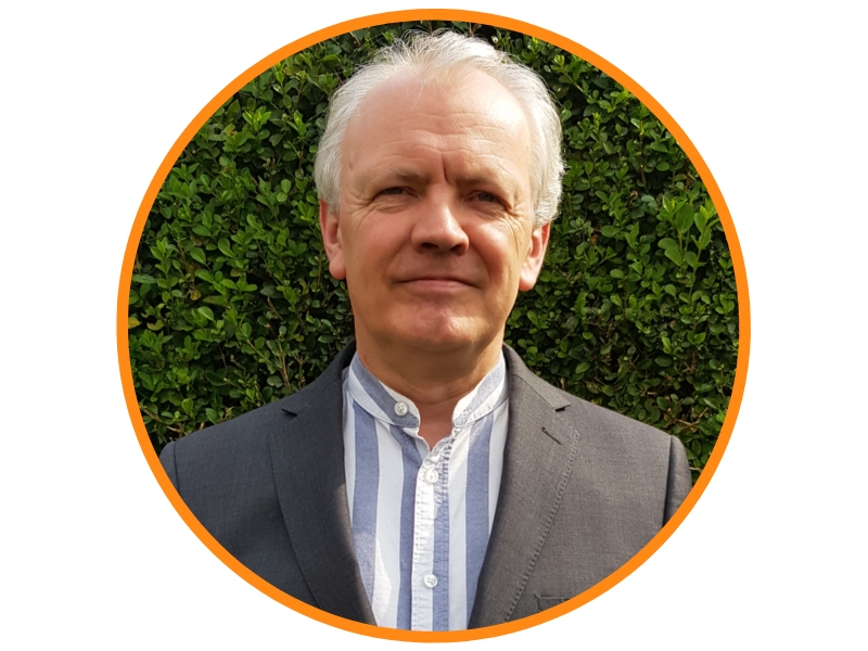 Man smiling and standing in front of a green hedge. Wearing a blue and white striped shirt with a grey blazer.